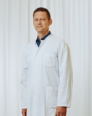 Ein Mann in einem weißen Laborkittel steht aufrecht vor einem hellen, neutralen Hintergrund.