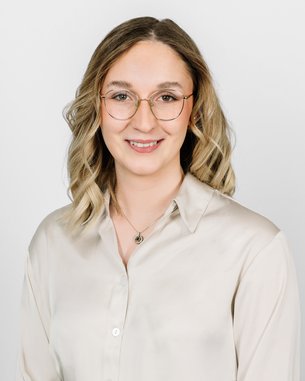 Eine Person mit blonden, lockigen Haaren und Brille trägt ein helles Hemd. Der Hintergrund ist neutral und hell.