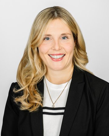 Eine lächelnde Frau mit langen, blonden Haaren trägt einen schwarzen Blazer und ein weißes Oberteil. Der Hintergrund ist neutral.