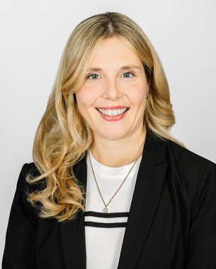 Eine lächelnde Frau mit langen, blonden Haaren trägt einen schwarzen Blazer und ein weißes Oberteil. Der Hintergrund ist neutral.