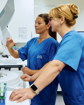 Zwei medizinische Fachkräfte in blauen Arbeitskleidung stehen an einem Laborgerät und betrachten den Bildschirm. Eine der Frauen zeigt auf den Bildschirm, während die andere aufmerksam zuschaut. Im Vordergrund sind Probenröhrchen auf einem Gestell sichtbar.