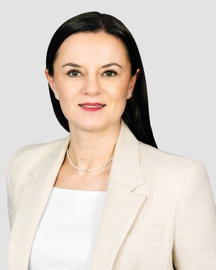 Eine Frau mit langen, dunklen Haaren trägt einen hellen Blazer und schaut direkt in die Kamera. Der Hintergrund ist neutral.