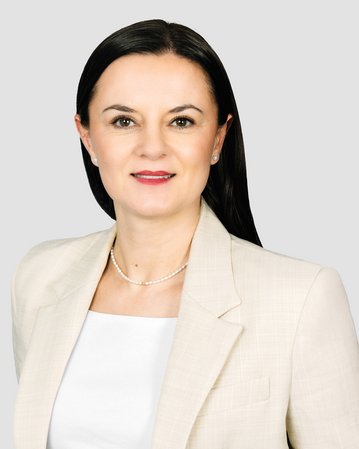 Eine Frau mit langen, dunklen Haaren trägt einen hellen Blazer und schaut direkt in die Kamera. Der Hintergrund ist neutral.