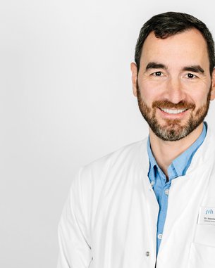 Ein Arzt mit Bart trägt einen weißen Kittel und lächelt. Er steht vor einem neutralen Hintergrund und hat ein Namensschild an.