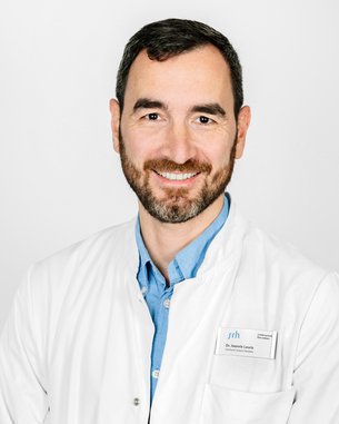 Ein Arzt mit Bart trägt einen weißen Kittel und lächelt. Er steht vor einem neutralen Hintergrund und hat ein Namensschild an.