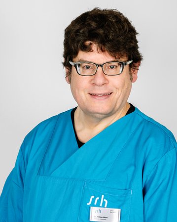 Ein Mann in einem blauen medizinischen Kittel steht vor einem neutralen Hintergrund. Er trägt eine Namensschilder.