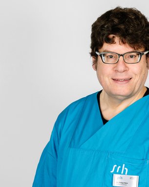 Ein Mann in einem blauen medizinischen Kittel steht vor einem neutralen Hintergrund. Er trägt eine Namensschilder.