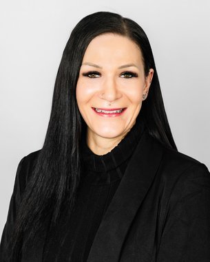 Eine Frau mit langen, schwarzen Haaren und einem schwarzen Blazer lächelt in die Kamera vor einem neutralen Hintergrund.