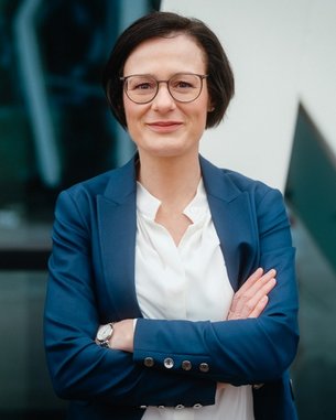 Frau mit kurzen, dunklen Haaren und Brille, gekleidet in einem blauen Blazer, steht mit verschränkten Armen vor einem modernen Gebäude.