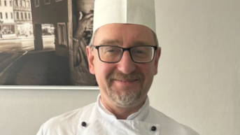 Ein Koch in einer weißen Uniform mit einer Kochmütze steht lächelnd vor einer Wand mit einem Schwarz-Weiß-Bild im Hintergrund.