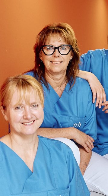 Fünf Frauen in blauen Arbeitskleidung stehen und sitzen in einem Flur. Sie lächeln und posieren freundlich für die Kamera. Der Hintergrund zeigt eine orangefarbene Wand und einen hellen Flur.