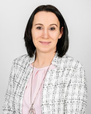 Eine Frau mit schulterlangen, dunklen Haaren trägt eine Bluse und eine Jacke mit Karomuster. Sie lächelt leicht vor einem neutralen Hintergrund.