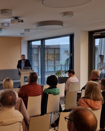 Ein Mann steht an einem Rednerpult und präsentiert vor einem Publikum in einem modernen Raum. Auf einer Leinwand sind Informationen zu sehen, während die Zuhörer aufmerksam zuhören.