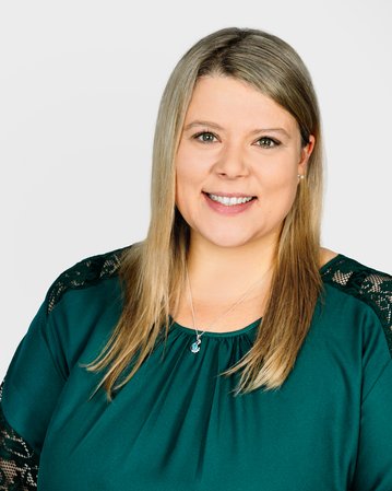 Eine lächelnde Frau mit langen, blonden Haaren trägt ein grünes Oberteil mit schwarzen Spitzenärmeln. Der Hintergrund ist neutral.