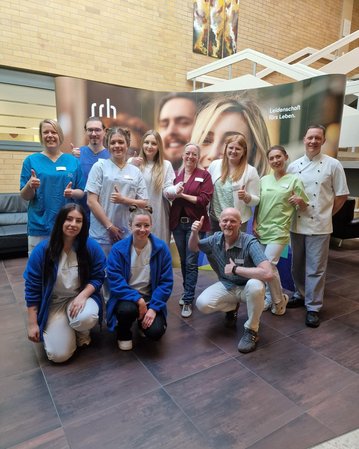 Eine Gruppe von zwölf Personen in Arbeitskleidung steht in einem Innenraum. Sie lächeln und zeigen Daumen hoch. Im Hintergrund ist ein Banner mit dem Text "Leidenschaft fürs Leben" sichtbar.