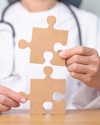 Eine Person in einem weißen Kittel hält zwei Puzzlestücke aus Karton, die sie zusammenfügt. Ein Stethoskop ist um den Hals der Person drapiert.