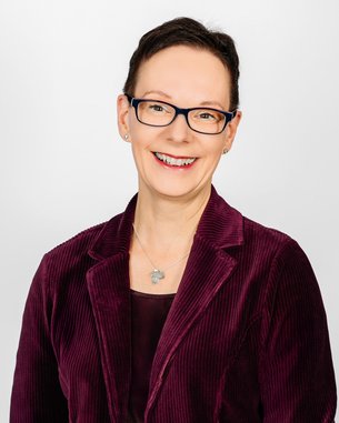 Eine lächelnde Frau mit Brille trägt einen lila Cord-Blazer. Der Hintergrund ist neutral und hell.