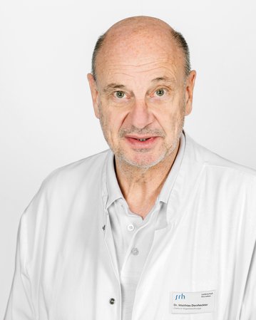 Ein Arzt in einem weißen Kittel steht vor einem neutralen Hintergrund und schaut direkt in die Kamera. Ein Namensschild ist sichtbar.