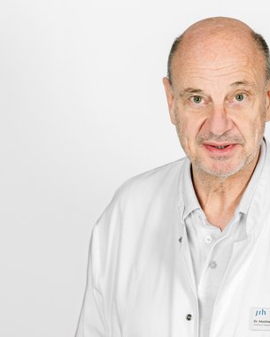 Ein Arzt in einem weißen Kittel steht vor einem neutralen Hintergrund und schaut direkt in die Kamera. Ein Namensschild ist sichtbar.