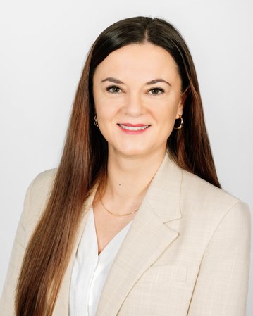 Eine Frau mit langen, braunen Haaren trägt einen hellen Blazer und lächelt in die Kamera. Der Hintergrund ist neutral und hell.