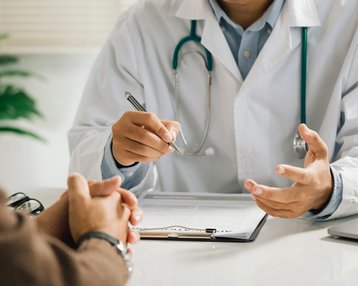 Ein Arzt in einem weißen Kittel erklärt etwas, während er mit einem Stift auf ein Dokument zeigt. Ein Patient sitzt gegenüber und hat die Hände gefaltet. Ein Stethoskop hängt um den Hals des Arztes.