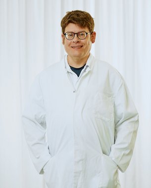 Eine Person in einem weißen Laborkittel steht mit den Händen in den Taschen vor einem hellen, neutralen Hintergrund. Die Person trägt eine Brille und hat kurzes, braunes Haar.