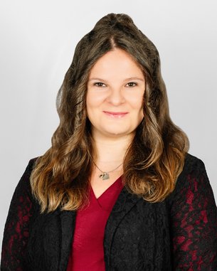 Eine lächelnde Frau mit langen, lockigen Haaren trägt eine schwarze Jacke über einem roten Oberteil. Der Hintergrund ist neutral.