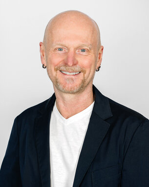 Mann mit Glatze und Ohrstecker, trägt einen schwarzen Blazer über einem weißen T-Shirt, lächelt freundlich vor neutralem Hintergrund.