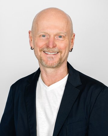 Mann mit Glatze und Ohrstecker, trägt einen schwarzen Blazer über einem weißen T-Shirt, lächelt freundlich vor neutralem Hintergrund.