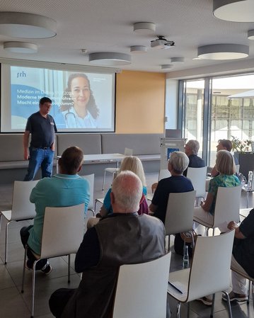 Ein Referent steht vor einem Publikum in einem modernen Raum. Auf einer Leinwand ist eine Präsentation mit dem Titel "Medizin im Jahr 2023" und einem Bild einer Frau mit einem Stethoskop zu sehen.