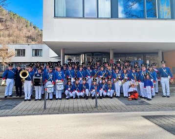 Eine große Gruppe von Musikern in Uniform steht vor einem modernen Gebäude. Die Mitglieder halten verschiedene Musikinstrumente, darunter Trompeten und Trommeln, und posieren für ein Gruppenfoto.