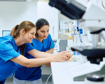 Zwei Frauen in blauen Laborkitteln betrachten gemeinsam ein Probenbehältnis an einem Labortisch, umgeben von wissenschaftlichen Geräten und Materialien.
